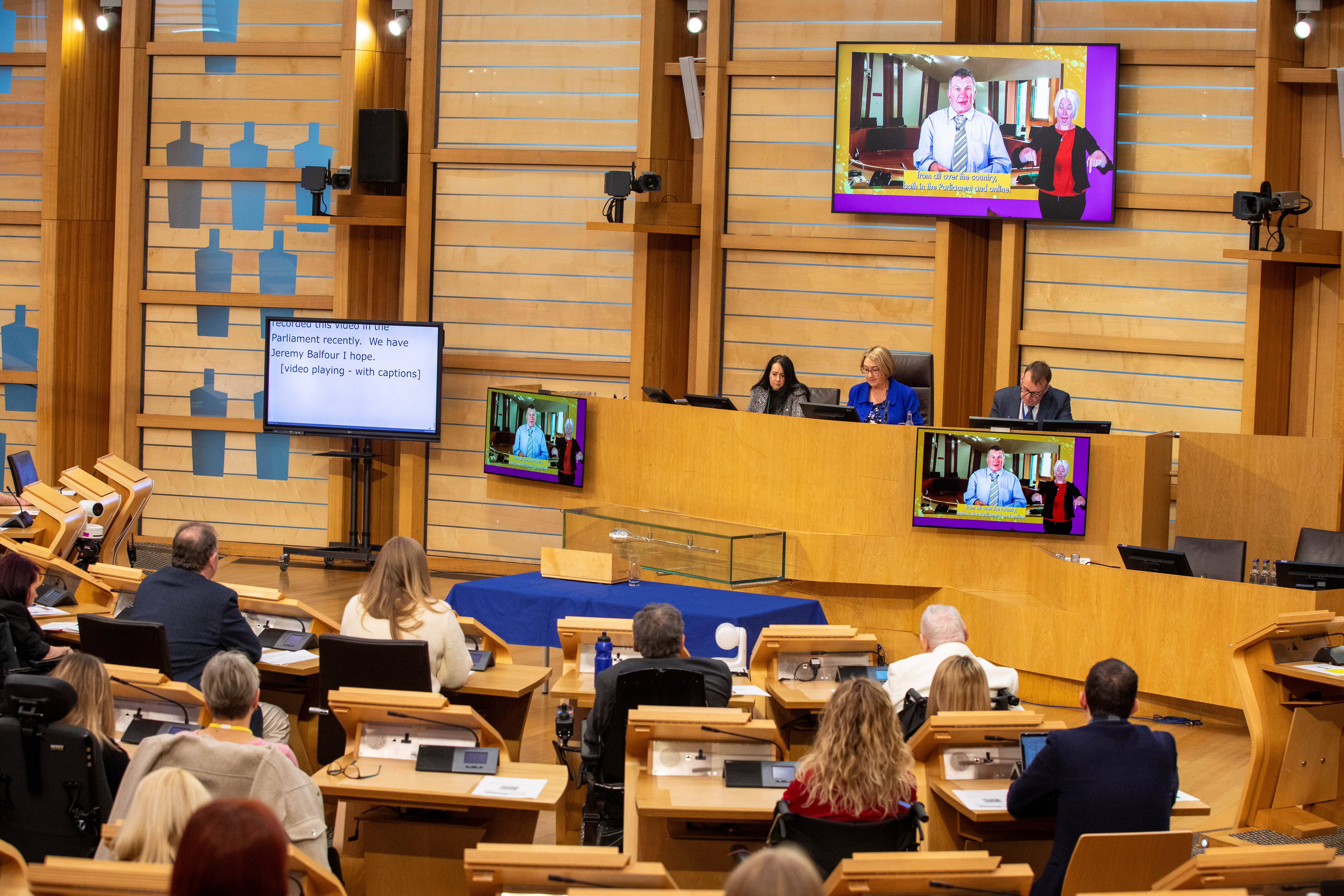 People sit at desks watching a man speaking on several screens. A sign language interpreter on the screen provides live interpretation. A separate screen provides a live transcript.